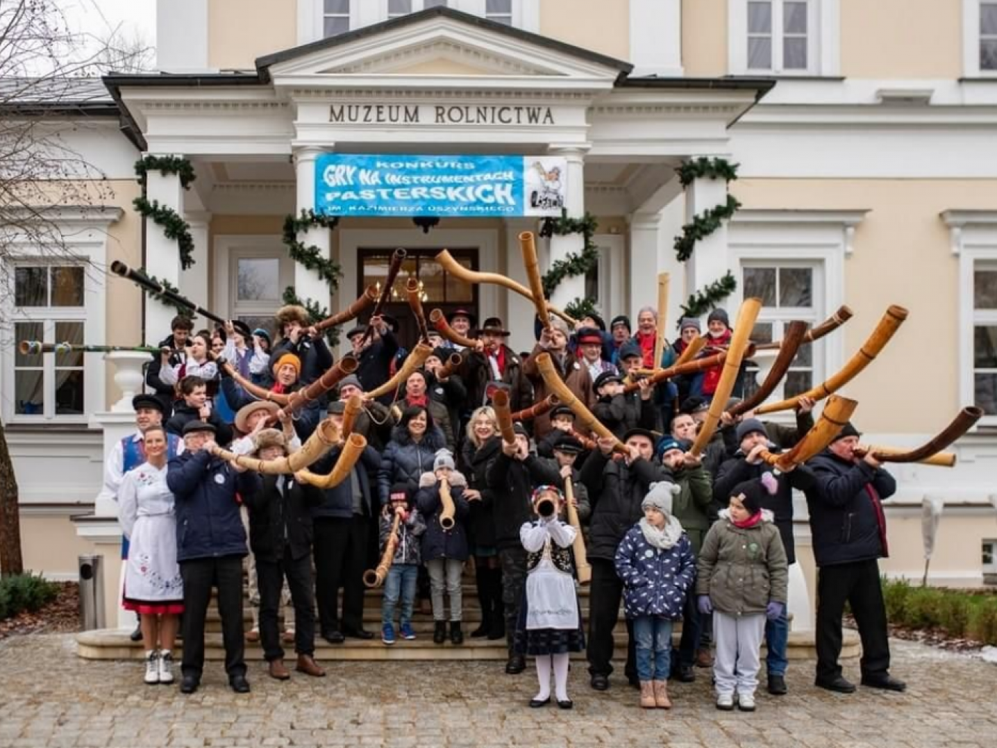 Group of people standing on steps in front of a building blowing various horn instruments.