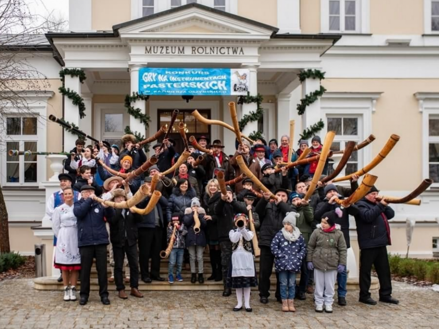 Group of people standing on steps in front of a building blowing various horn instruments. 