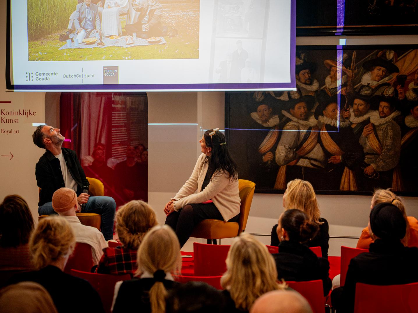 Panelleden en publiek kijken naar beeld op scherm