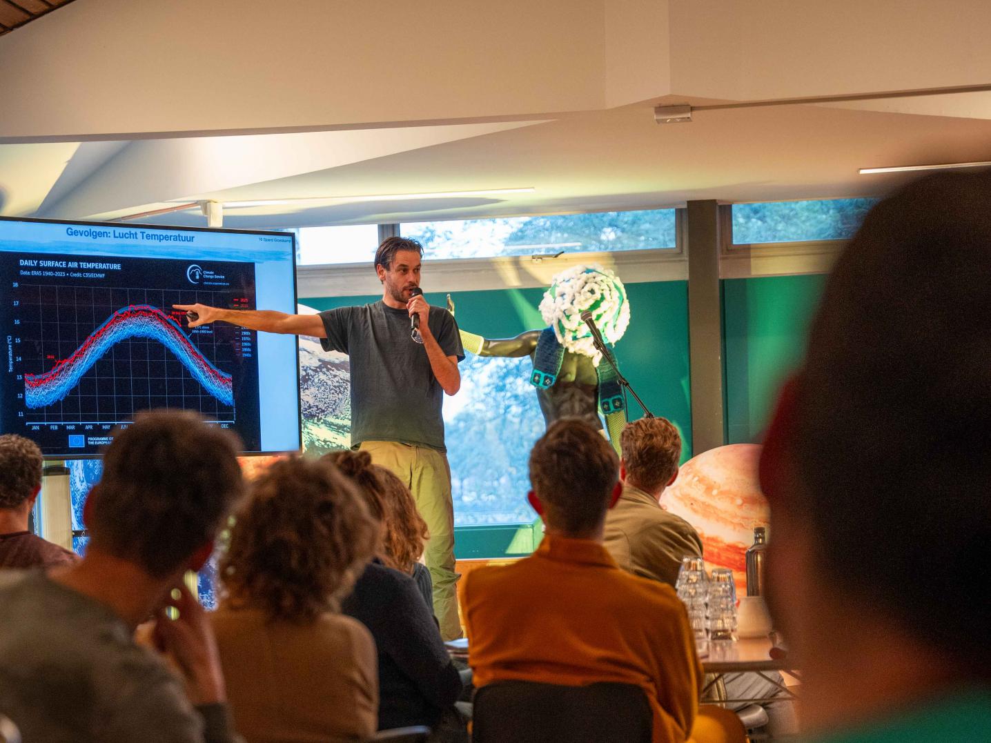 a room full pf people sitting and listening to a man speaking and pointing to a screen depicting a chart