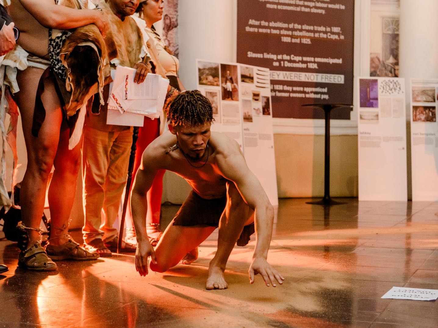A dancer crouched on the floor, engaging in a performance. The public is gathered around, close to the performer. surroundings are made up of informative museum pannels and the public is