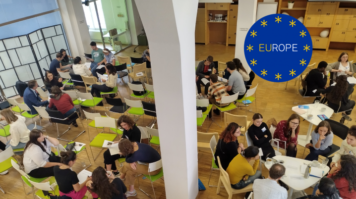 Overview of room full of chairs, people sit in small groups at tables discussing